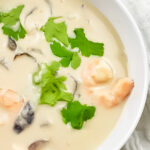 overhead zoomed in pic of tom kha goong in white bowl surrounded by white cloth.