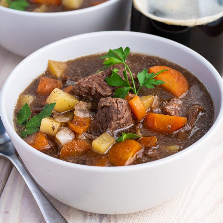 Gluten Free Slow Cooker Beef stew in a grey bowl next to a glass of stout beer, another bowl of beef stew, and a spoon.