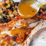 Gravy being spooned over turkey on a white plate with brussels sprouts and stuffing.