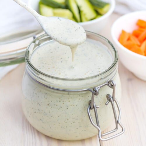 A spoon drizzles Whole30 ranch dressing into a glass jar with a metal clasp on a light-colored surface. In the background are bowls of carrot sticks and cucumber slices.