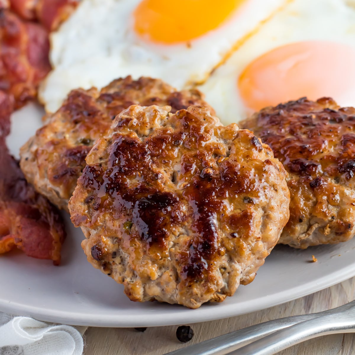 Three cooked keto breakfast sausage patties on a plate with bacon strips and two sunny-side-up eggs in the background.