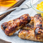 Air Fryer sausages on a baking tray with a fork, a glass of beer and bowl of mustard.