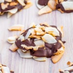 A cluster of coconut clusters, with toasted flakes partially coated in dark chocolate, sits on a light wooden surface.