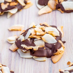 A cluster of coconut clusters, with toasted flakes partially coated in dark chocolate, sits on a light wooden surface.