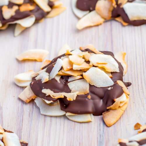 A cluster of coconut clusters, with toasted flakes partially coated in dark chocolate, sits on a light wooden surface.