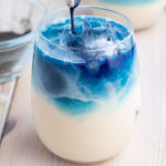 Close-up of a layered Butterfly Pea Milk Tea in a clear glass, featuring a blue liquid poured over ice cubes on top of a creamy white base. The drink has a visually striking gradient from blue at the top to white at the bottom. A blurred bowl is in the background.