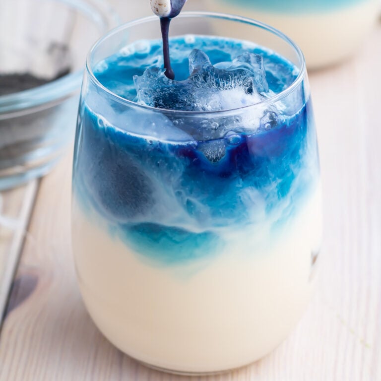 Close-up of a layered Butterfly Pea Milk Tea in a clear glass, featuring a blue liquid poured over ice cubes on top of a creamy white base. The drink has a visually striking gradient from blue at the top to white at the bottom. A blurred bowl is in the background.