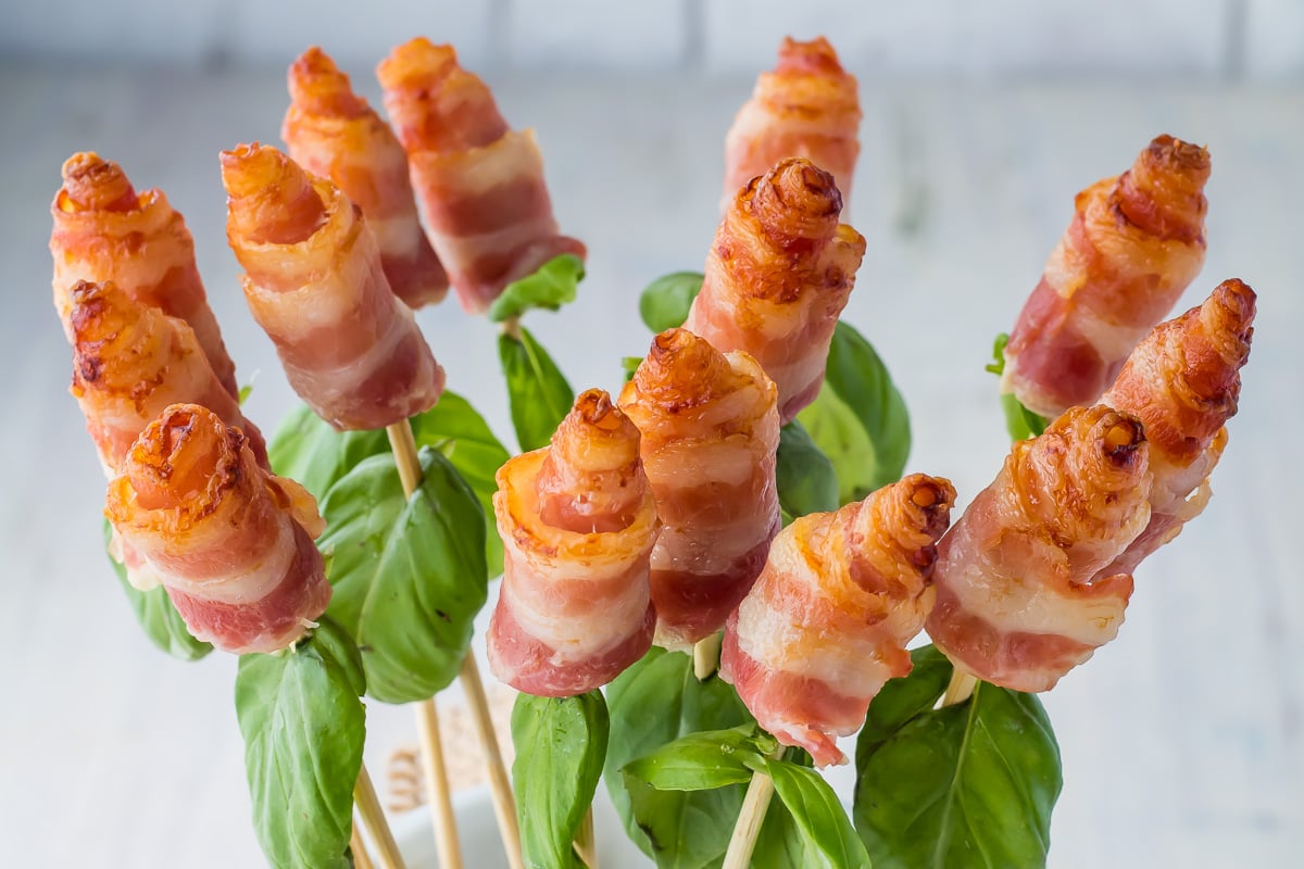 A bouquet of Bacon Roses on skewers is shown. Each skewer has a rolled bacon slice resembling a rosebud, arranged on a base of fresh basil leaves. The background is light and unobtrusive, highlighting the detailed texture and color of the bacon.