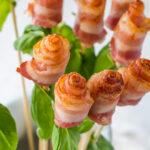 Bacon Roses on skewers, garnished with fresh green basil leaves. The bacon is cooked to a golden brown, tightly rolled to resemble rosebuds, and arranged artfully. The background is a subtle, light surface that complements the dish perfectly.