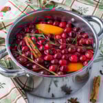 Christmas simmer pot ingredients in a pot next to a Christmas tea towel, herbs, and spices.