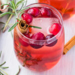 Two glasses of Christmas gin on a white table with rosemary, cinnamon stick, and a Christmas tea towel.