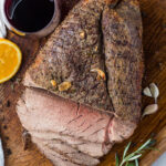 Overhead shot of sliced roast beef on a wooden cutting board next to wine, rosemary, and an orange.