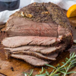 Sliced roast beef on a wooden cutting board next to wine, rosemary, and an orange.