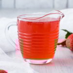 A jug of strawberry syrup on a table with strawberries and a white cloth.