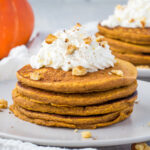 A stack of paleo pumpkin pancakes on a grey plate with coconut whipped cream and walnuts on top.