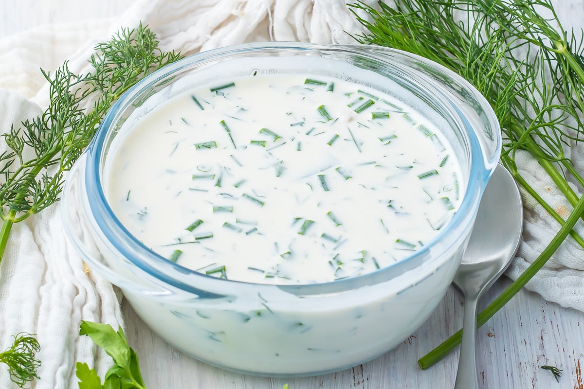 A bowl of keto ranch dressing next to fresh dill, on a white wooden surface with a spoon at the side.