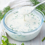 A bowl of creamy keto ranch dressing with a spoon drizzling some, surrounded by fresh dill and parsley on a wooden table.