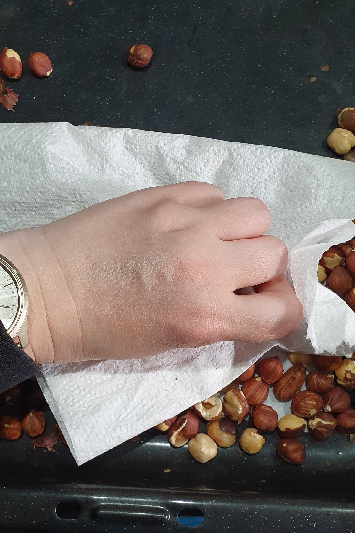 A person's hand with a wristwatch placing hazelnuts on a paper towel on a dark surface, demonstrating how to roast hazelnuts.