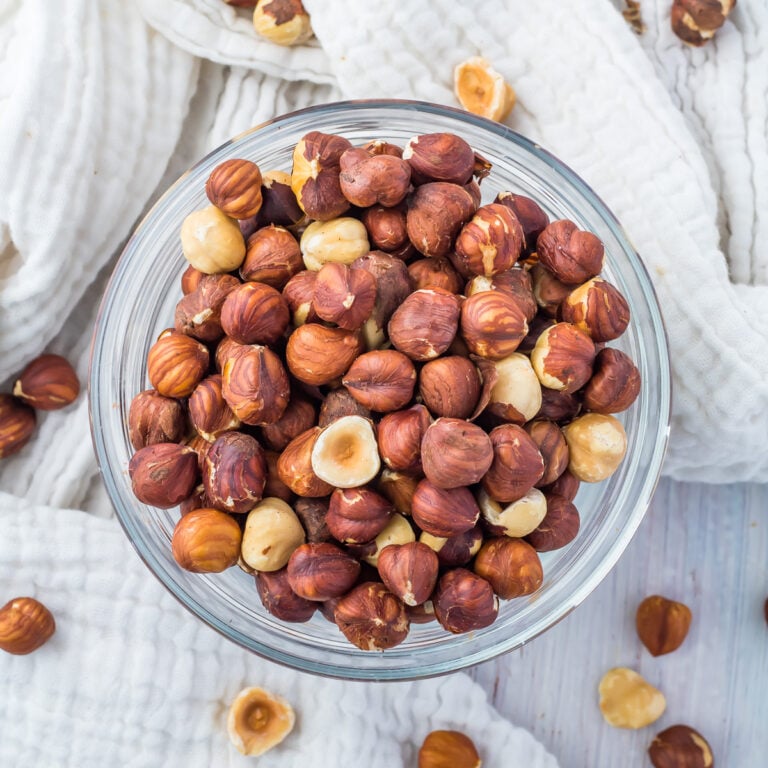 A glass bowl filled with whole and halved hazelnuts, placed on a white cloth with loose nuts scattered around, ready to demonstrate how to roast hazelnuts.