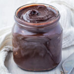 A glass jar filled with sugar free glossy dark Nutella spread on a wooden table, accompanied by a metal spoon and a white cloth.
