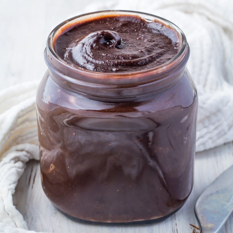A glass jar filled with sugar free glossy dark Nutella spread on a wooden table, accompanied by a metal spoon and a white cloth.