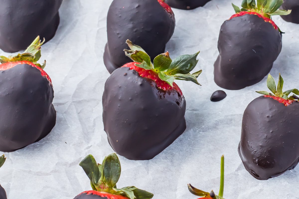 Close-up of fresh strawberries dipped in dark chocolate and Nutella, arranged on a parchment-lined tray with some chocolate drips visible.