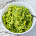 A white bowl filled with mashed keto guacamole, showing a mix of green and yellow hues with pieces of herbs visible. A spoon rests on the edge of the bowl.