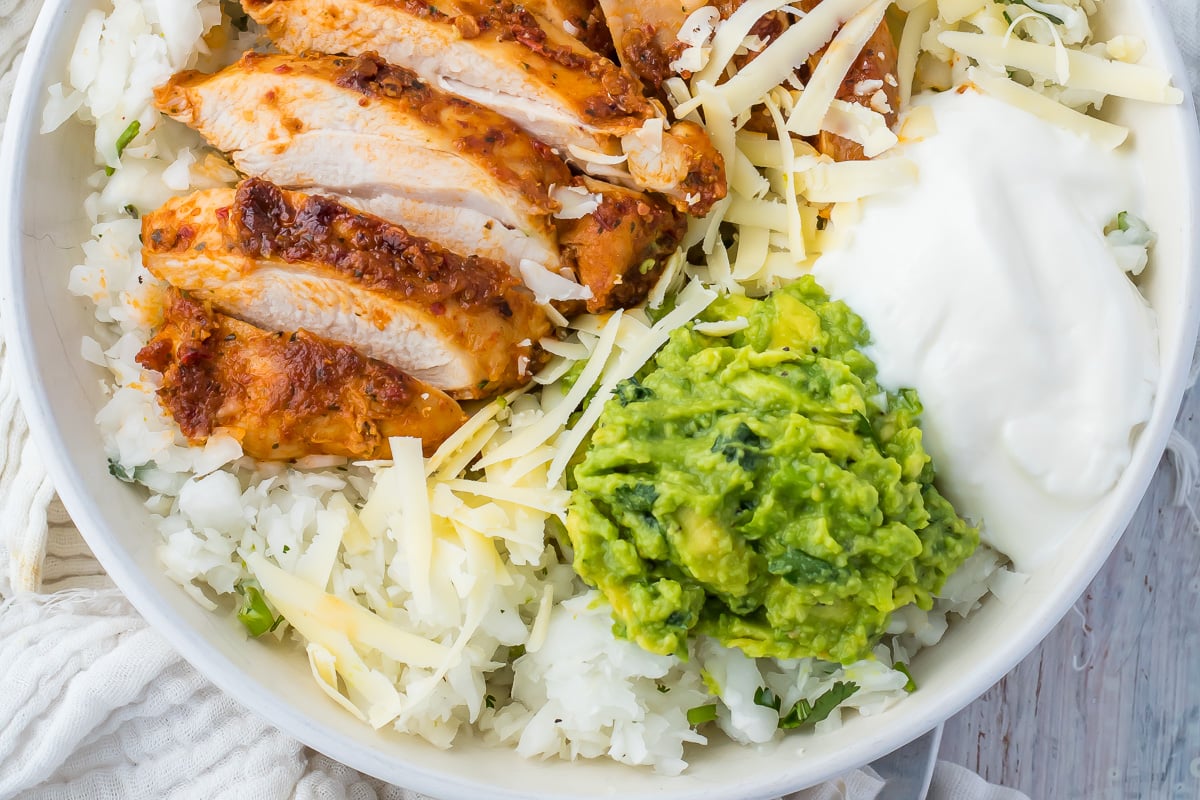 A close-up of a dish featuring sliced grilled chicken, shredded cheese, a dollop of sour cream, keto guacamole, and white radish rice on a plate.