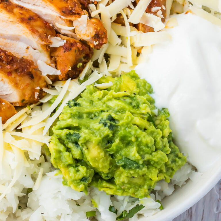 A close-up photo shows a dish with shredded chicken, grated cheese, keto guacamole, and a dollop of sour cream on top of a bed of white radish rice.