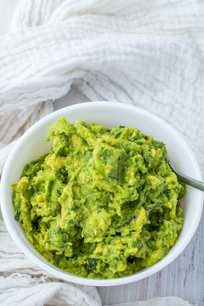 A bowl filled with mashed keto guacamole, placed on a wooden surface with a white fabric in the background.