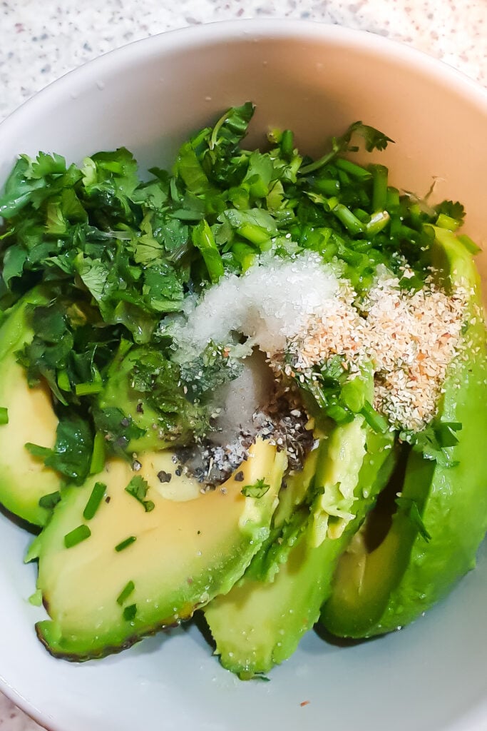 A bowl containing sliced avocado, chopped cilantro, and various seasonings including salt and pepper.