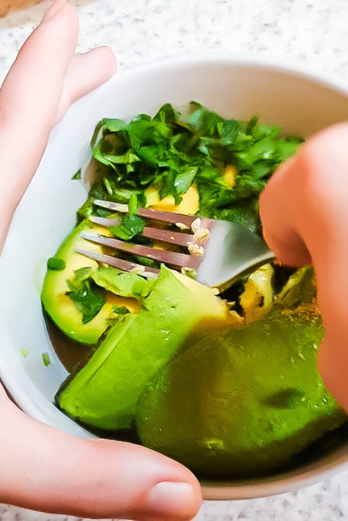 A person mashes avocado and green herbs with a fork in a white bowl.