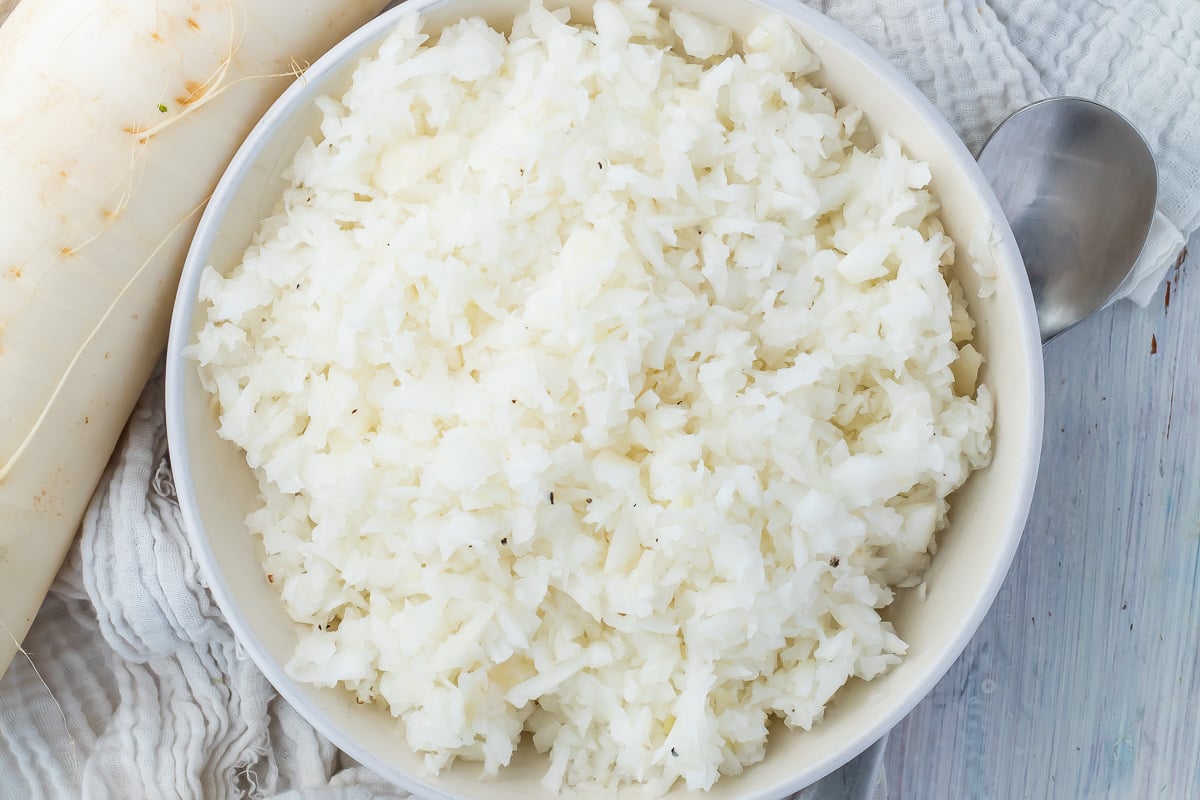 A bowl of cooked radish rice on a wooden table, with a spoon and daikon radish to the side.