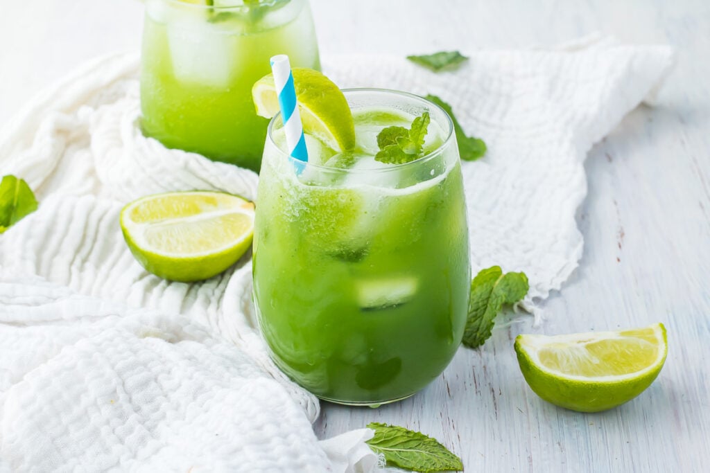 A refreshing cucumber cooler with ice cubes, garnished with a lime slice and mint leaves, is served in a glass with a blue and white striped straw. Additional lime slices and mint leaves are scattered around.