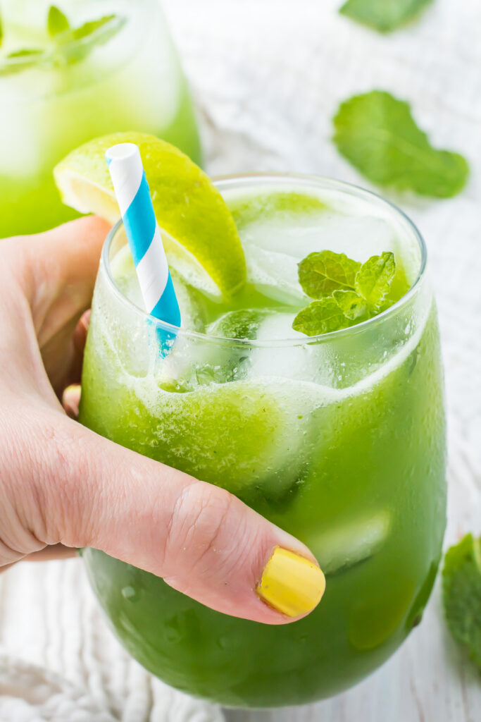 A hand with yellow nail polish holds a glass of cucumber cooler with ice, mint leaves, a lime slice, and a blue and white striped straw. Another similar glass is blurred in the background.