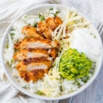 A keto burrito bowl of sliced grilled chicken on a bed of radish rice, topped with shredded cheese, guacamole, and sour cream.
