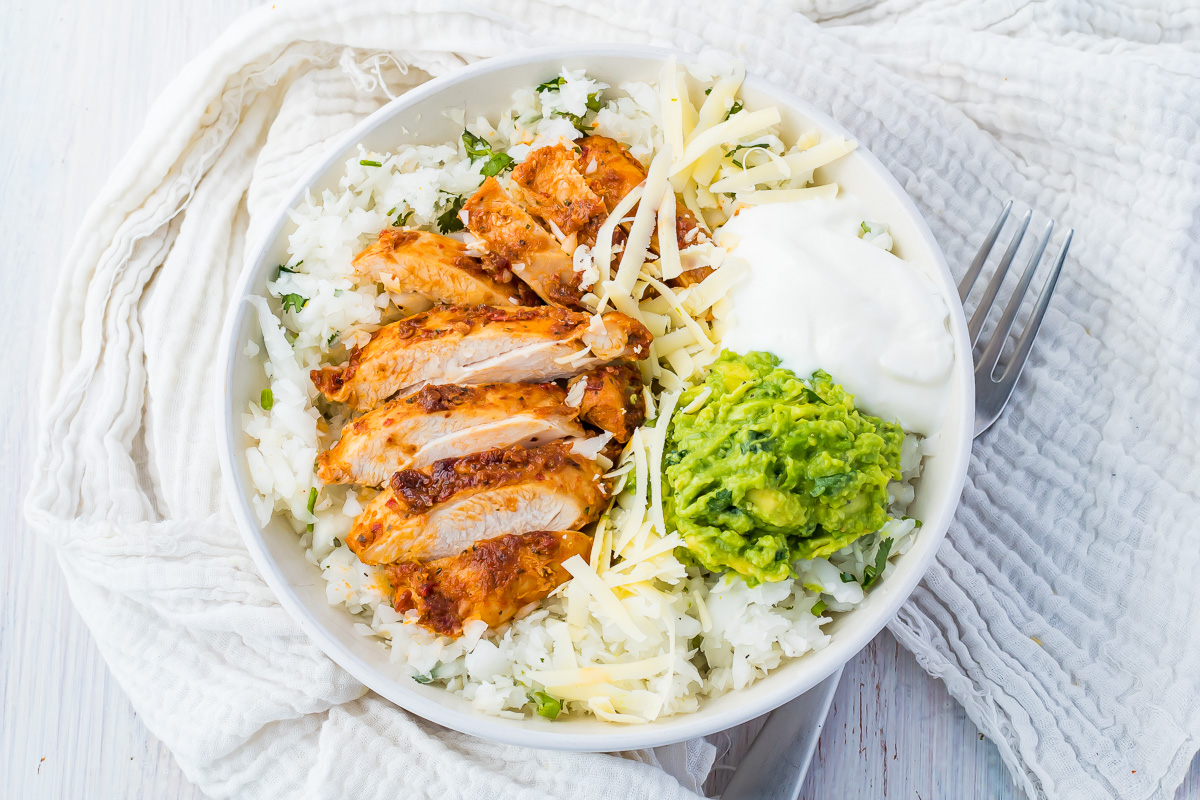 A keto chipotle bowl of radish rice topped with slices of grilled chicken, shredded cheese, guacamole, and a dollop of sour cream, with a fork placed beside the bowl.