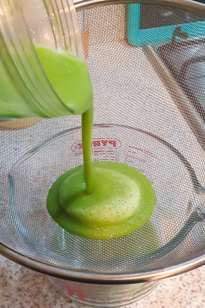 A refreshing cucumber cooler is being poured from a blender into a metal strainer over a measuring cup on the kitchen countertop.