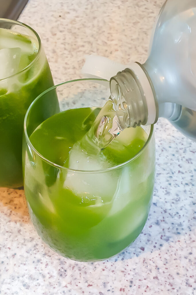 A bottle is pouring a club soda into a glass filled with cucumber cooler and ice cubes. Another glass with green juice and ice cubes is partially visible in the background.