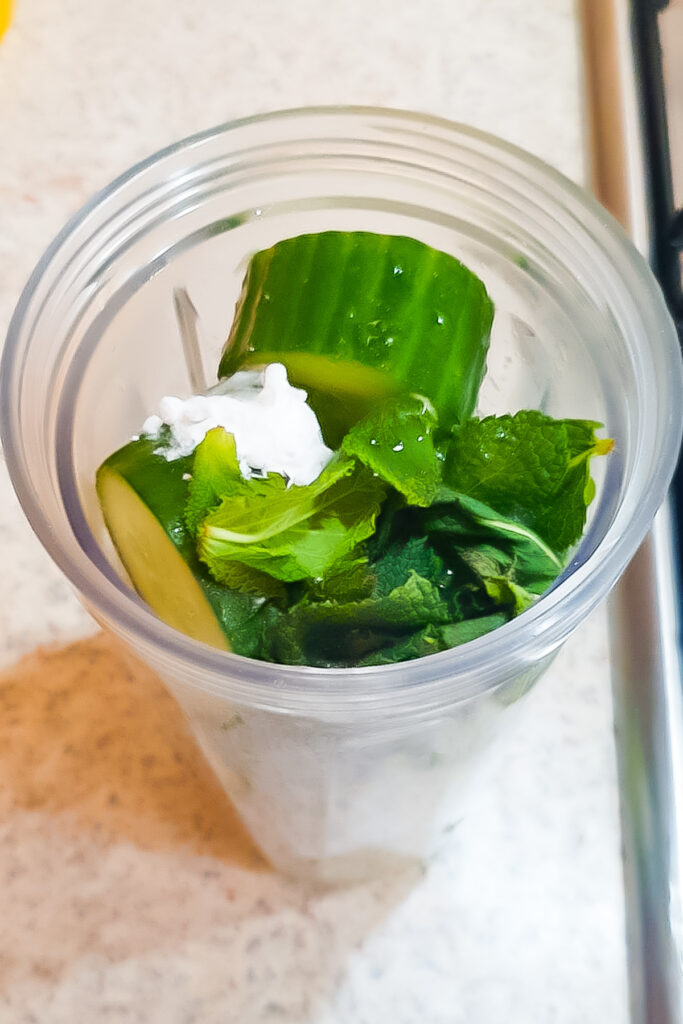 A plastic blender cup on a countertop contains sliced cucumber, fresh mint leaves, and a dollop of yogurt, ready to be transformed into a refreshing cucumber cooler.