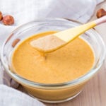 A glass bowl filled with creamy peanut butter, with a spoon partially dipped in and a few nuts scattered around on a white cloth background.