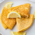 Three folded keto crepes topped with lemon slices and lightly dusted with sweetener, served on a white plate. The crepes have a golden brown color, and the lemon slices add a fresh contrast to the dish.