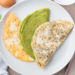 A white plate features three keto egg wraps: one orange, one green, and one speckled beige, arranged to form a semi-circle. Partial views of a brown egg, a white napkin, and other items are visible in the background.