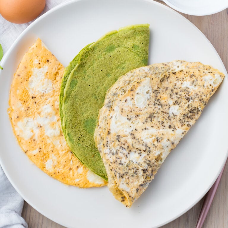 A white plate features three keto egg wraps: one orange, one green, and one speckled beige, arranged to form a semi-circle. Partial views of a brown egg, a white napkin, and other items are visible in the background.