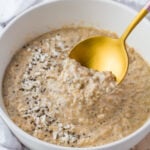 A close-up of a white bowl filled with creamy keto oatmeal topped with shredded coconut and chia seeds. A golden spoon is scooping some of the oatmeal, which appears to be cooked to a smooth, thick consistency.