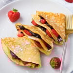 A white plate with two folded keto crepes filled with chocolate spread and sliced strawberries, dusted with powdered sugar. Fresh whole strawberries adorn the plate. A gold fork with a pink handle rests on the right side of the plate.