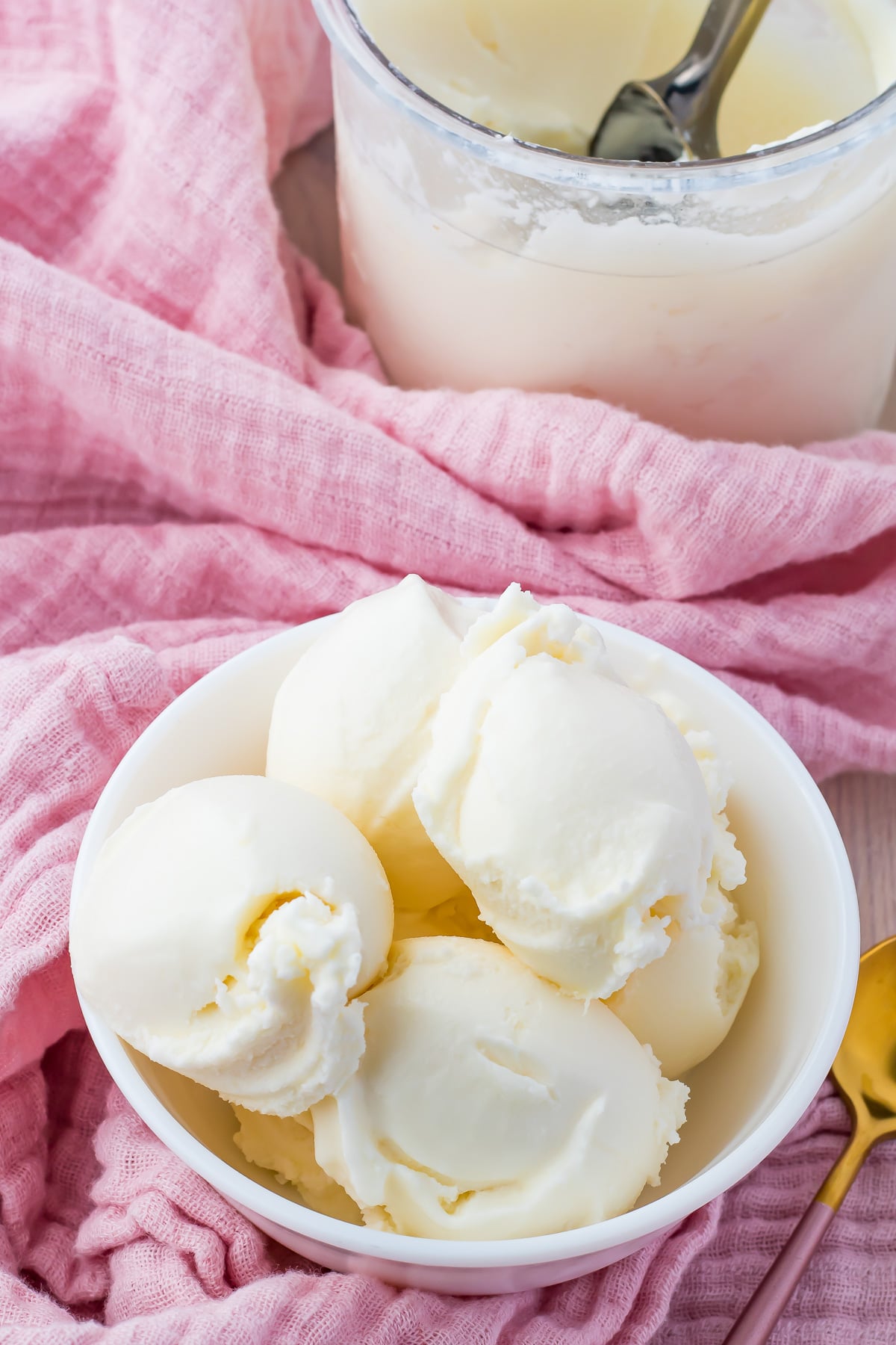 A bowl of creamy pineapple coconut ice cream with six scoops is placed on a light pink textured cloth. A container of ice cream with a spoon inside is partially visible in the background. A gold-colored spoon is also visible near the cloth.
