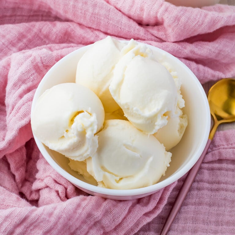 A white bowl filled with several scoops of pineapple coconut ice cream sits on a pink cloth. A gold spoon is placed beside the bowl on the cloth. The overall setting gives a cozy and inviting look.