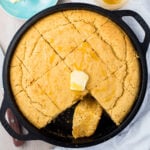A round skillet filled with golden-brown gluten-free cornbread is shown with a wedge cut out. The cornbread is topped with a pat of butter and drizzled with honey, and is placed on a light-colored surface.