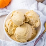 A bowl of creamy, keto pumpkin ice cream scoops, surrounded by a white cloth. A small pumpkin is partially visible in the background, and a gold spoon rests nearby.